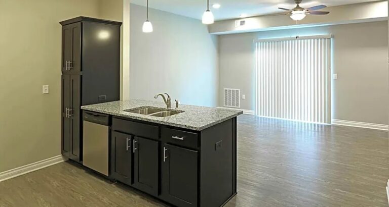 Modern kitchen with island sink, dark cabinets, dishwasher, hanging lights, and open living area with sliding door blinds.