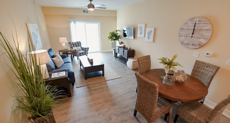 Open-plan living and dining area with neutral decor, wicker chairs, a wall clock, and large windows letting in natural light.