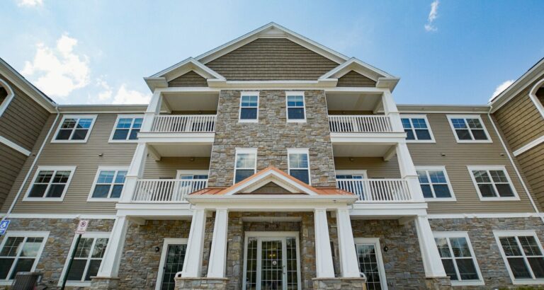 Three-story apartment building with stone and siding exterior, balconies, white trim, and a gabled roof under a blue sky.