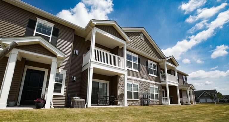 Two-story apartment building with balconies, brown siding, white trim, and a grassy lawn under a blue sky with clouds.