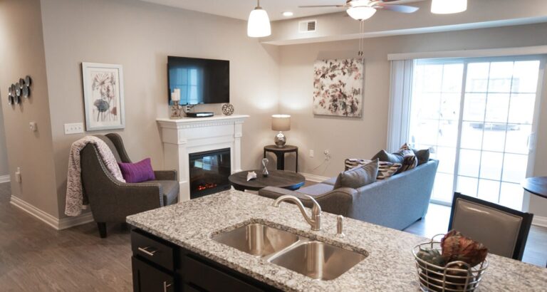 Modern living room with gray sofa, TV above a white fireplace, and sliding glass doors, viewed from a kitchen island.