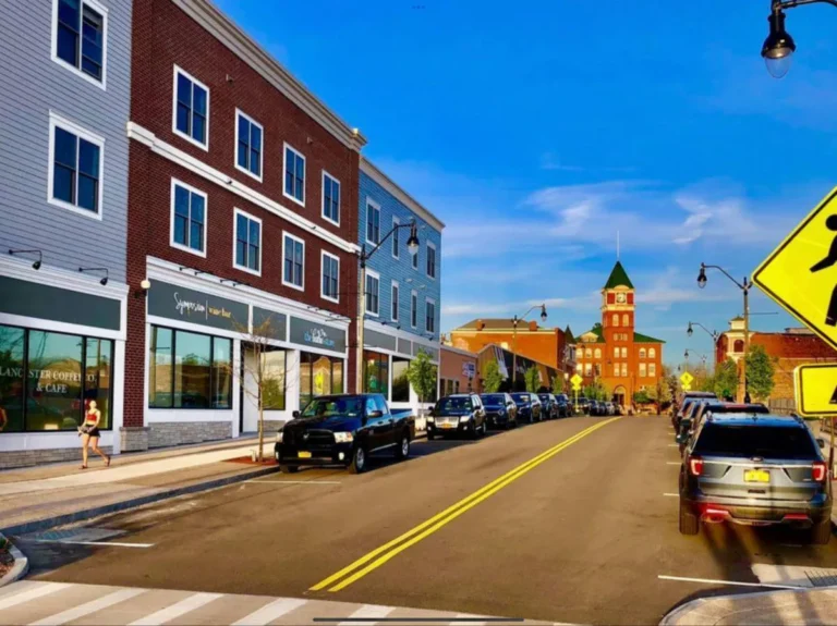 A downtown street with parked cars, storefronts on both sides, and a clock tower visible in the background under a blue sky.