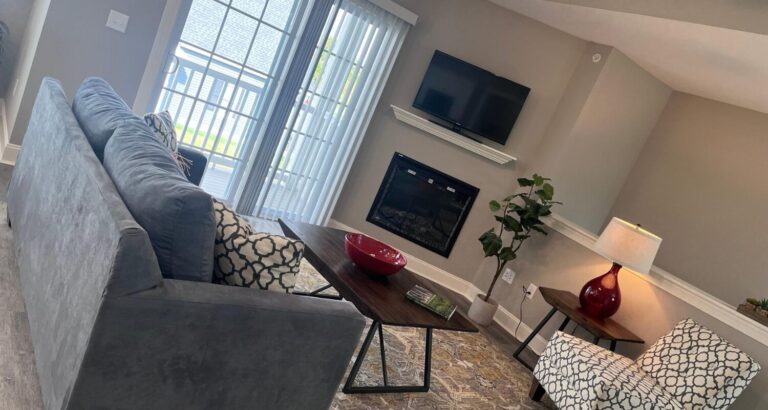 Modern living room with a gray sofa, patterned chairs, mounted TV, fireplace, and sliding glass doors to a balcony.