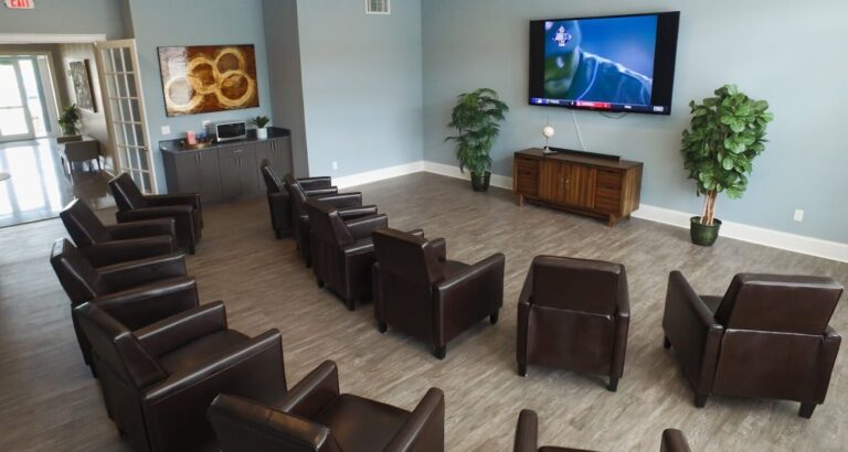 A lounge with brown leather chairs facing a large wall-mounted TV showing a sports game, with plants and a cabinet.