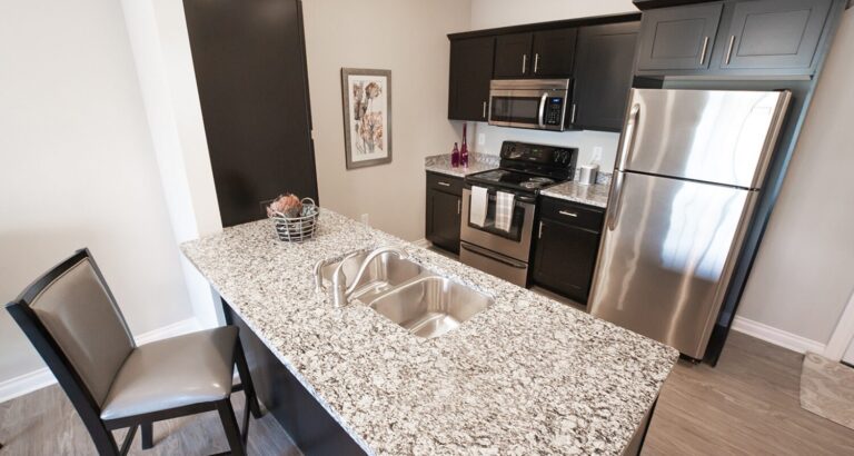 Modern kitchen with granite island, stainless steel appliances, dark cabinets, and a single chair by the counter.