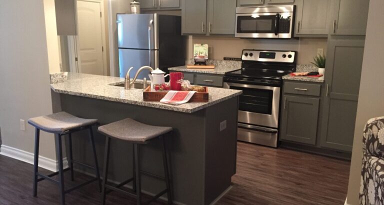 Modern kitchen with gray cabinets, stainless steel appliances, granite countertops, and two stools at a central island.